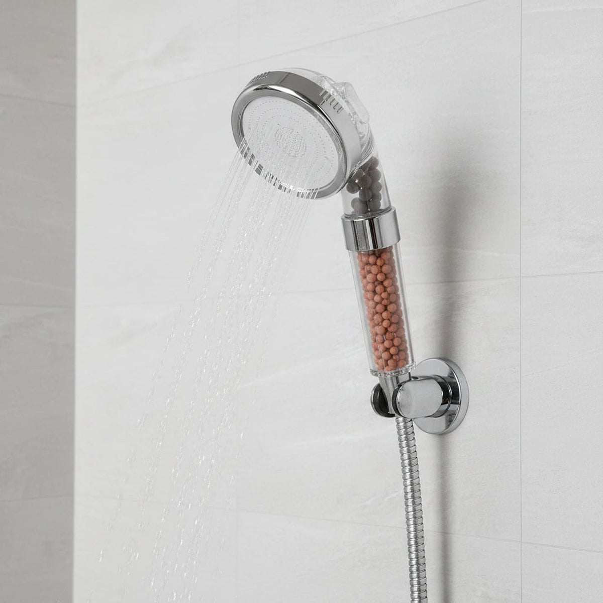 Filtered high pressure shower head mounted in a chrome wall bracket on a tiled shower wall, spraying clear pressurized water, with visible mineral filter beads in the clear handle